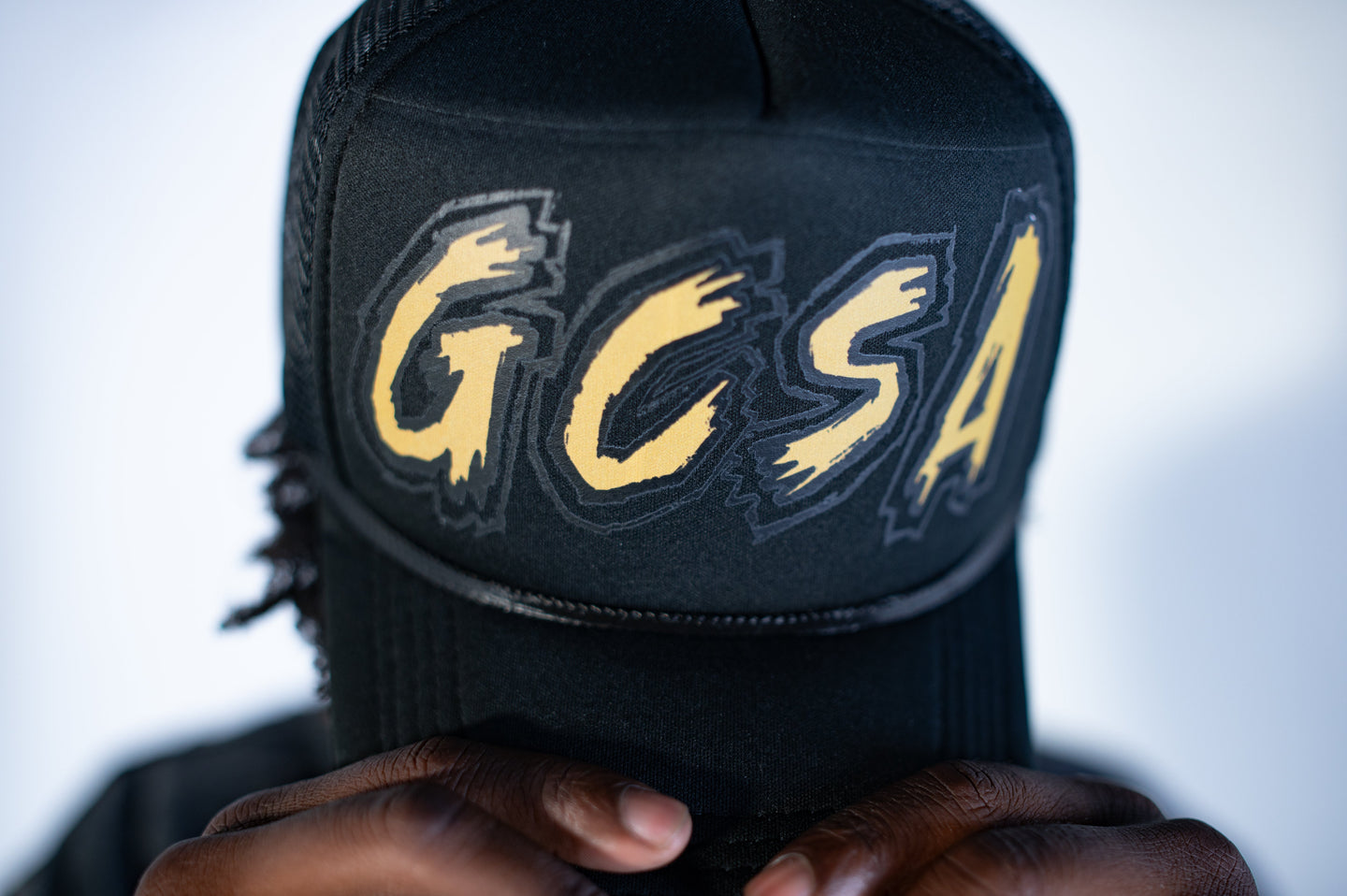 Black cap with 'GCSA' in gold letters held by a person against a white background