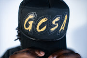 Black cap with 'GCSA' in gold letters held by a person against a white background
